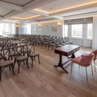 Elegant ceremony room at Buile Hill Mansion featuring wooden floors, rows of rustic chairs, a central ceremony table with two blush pink chairs, decorative wall panels, and warm ceiling lighting.
