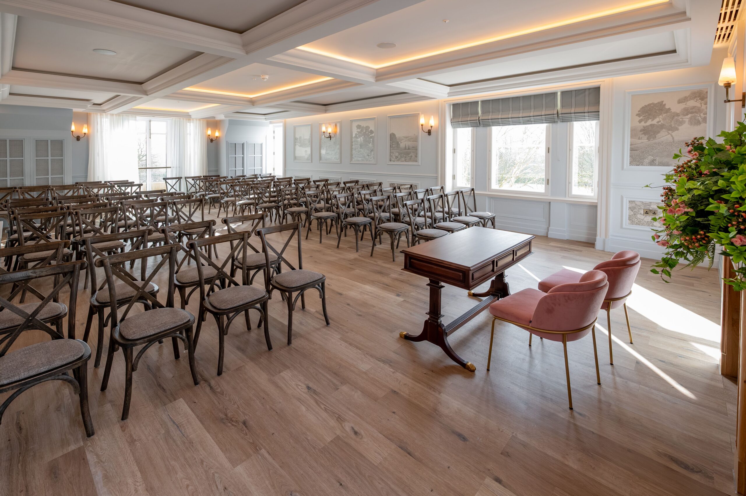 Elegant ceremony room at Buile Hill Mansion featuring wooden floors, rows of rustic chairs, a central ceremony table with two blush pink chairs, decorative wall panels, and warm ceiling lighting.