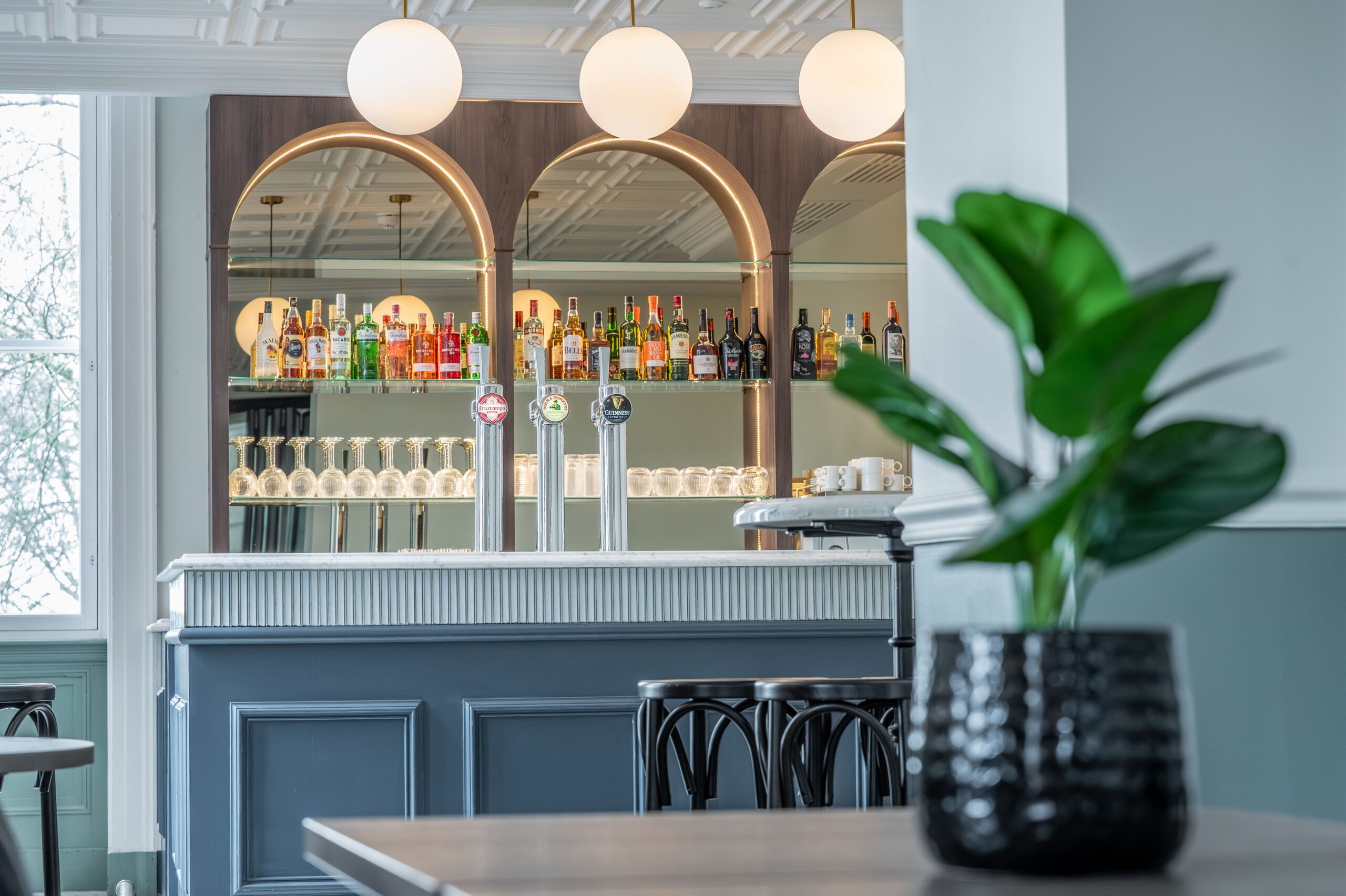 Contemporary bar area at Buile Hill Mansion featuring arched mirrored shelving with bottles and glassware, beer taps, soft globe pendant lights, and a classic panelled bar front.