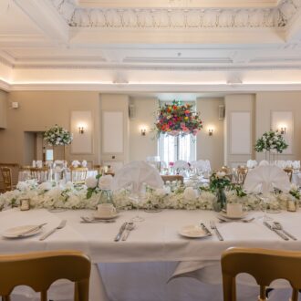Grand function room at Buile Hill Mansion featuring a floral-dressed top table, white table linens, gold chairs, tall floral centrepieces, chandeliers, and decorative ceiling detail.