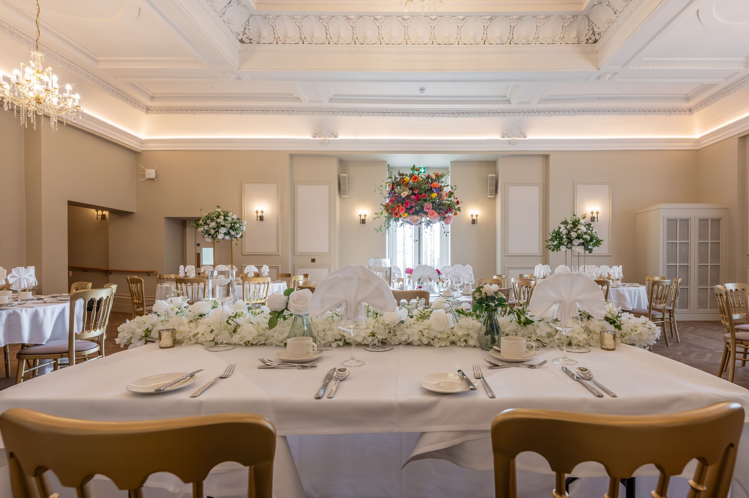 Grand function room at Buile Hill Mansion featuring a floral-dressed top table, white table linens, gold chairs, tall floral centrepieces, chandeliers, and decorative ceiling detail.