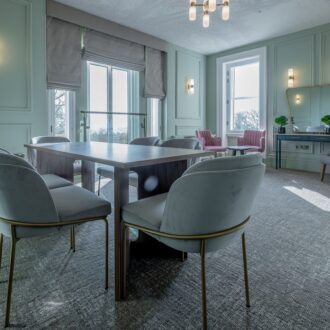 Light-filled bridal room at Buile Hill Mansion featuring a central table with soft grey chairs, panelled pale green walls, large sash windows, and modern wall and ceiling lights.