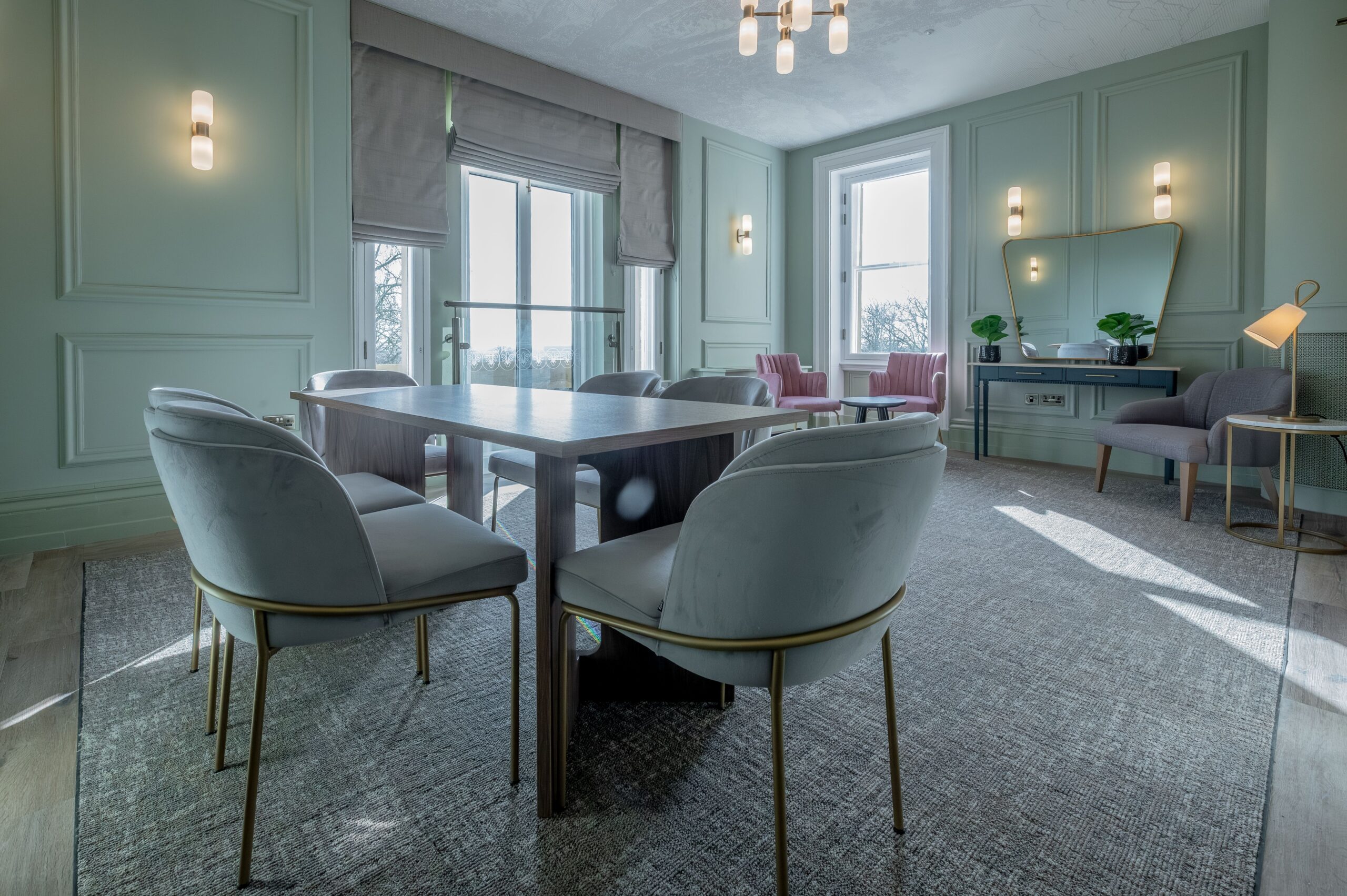 Light-filled bridal room at Buile Hill Mansion featuring a central table with soft grey chairs, panelled pale green walls, large sash windows, and modern wall and ceiling lights.