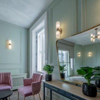 Elegant bridal room at Buile Hill Mansion featuring blush pink upholstered chairs, a console table with large mirror, potted plants, pale green panelled walls, and soft wall lighting beside tall windows.