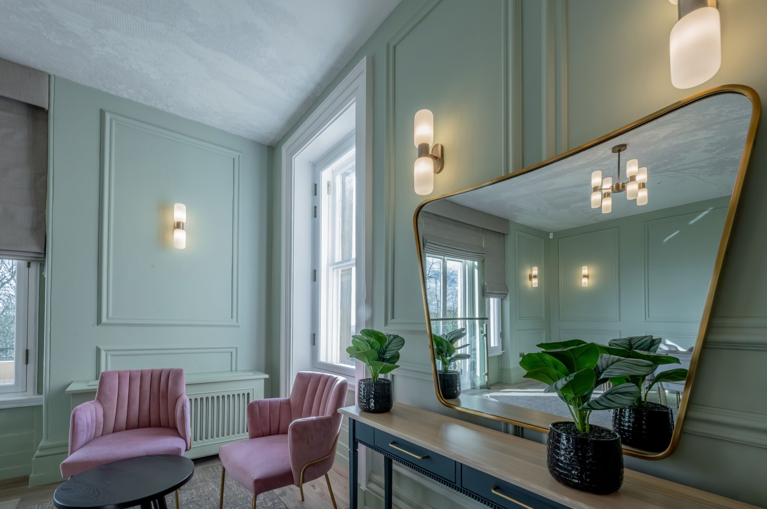 Elegant bridal room at Buile Hill Mansion featuring blush pink upholstered chairs, a console table with large mirror, potted plants, pale green panelled walls, and soft wall lighting beside tall windows.