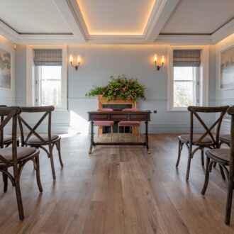 Elegant ceremony room at Buile Hill Mansion featuring wooden floors, rows of cross-back chairs, a central ceremony table with floral arrangement, pale blue panelled walls, and warm ceiling lighting.