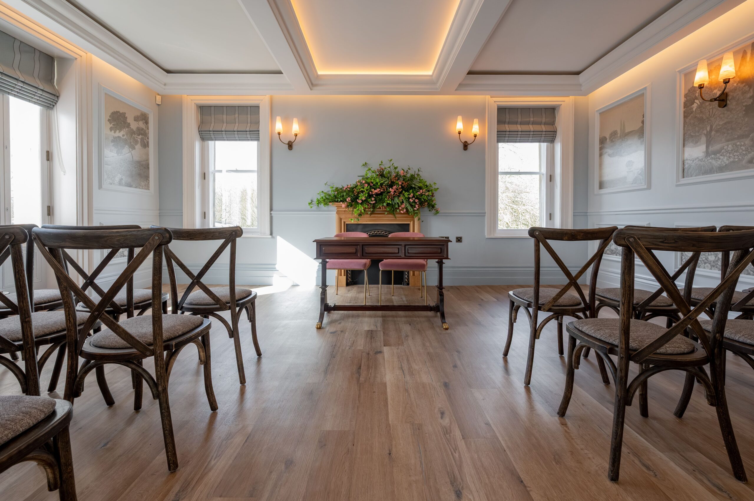 Elegant ceremony room at Buile Hill Mansion featuring wooden floors, rows of cross-back chairs, a central ceremony table with floral arrangement, pale blue panelled walls, and warm ceiling lighting.