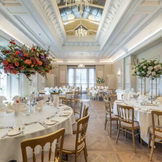 Grand reception room at Buile Hill Mansion with round tables laid with white tablecloths and folded napkins, gold chairs, tall floral displays, parquet flooring, and ornate ceiling details with chandeliers.