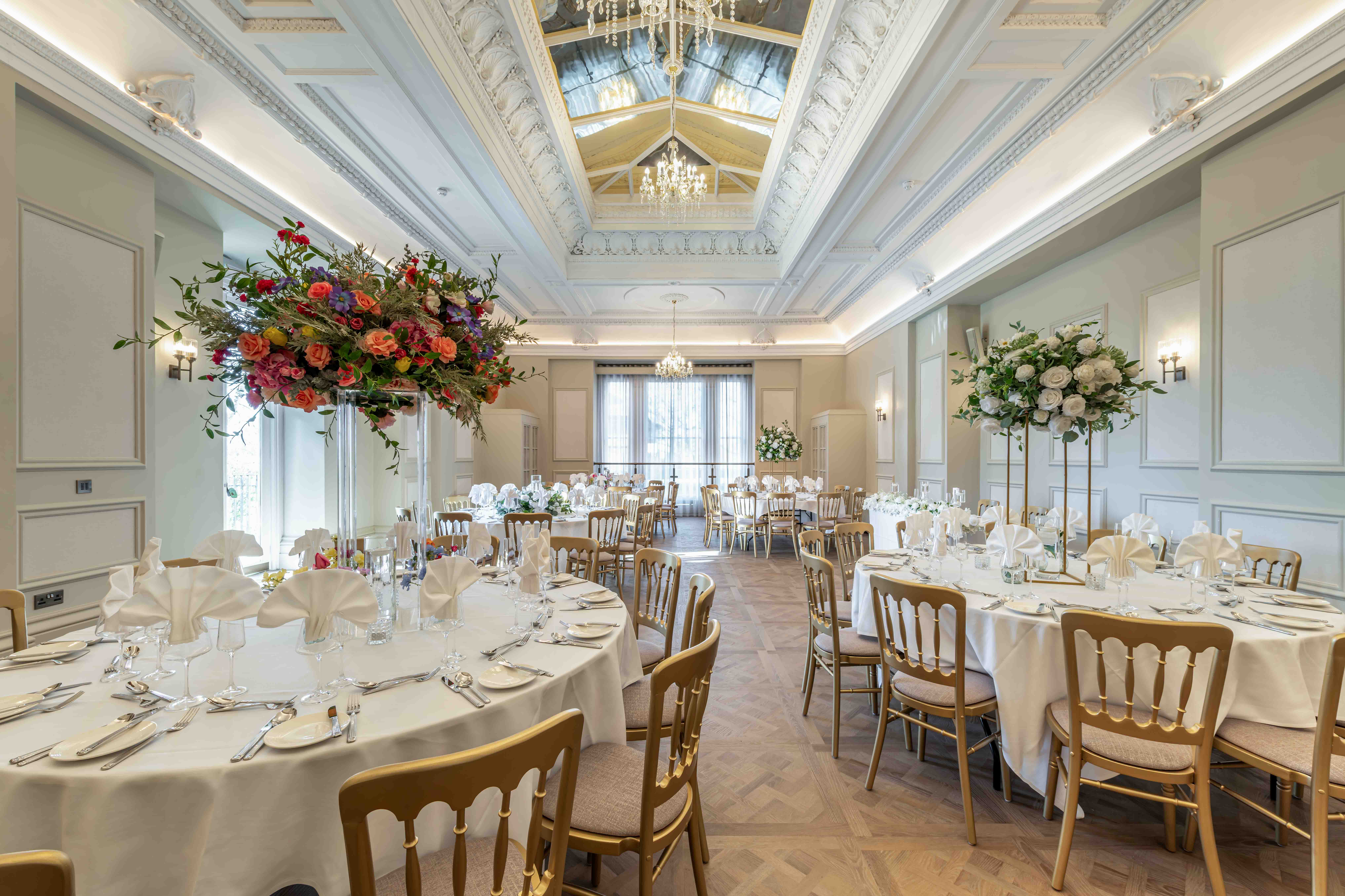 Grand reception room at Buile Hill Mansion with round tables laid with white tablecloths and folded napkins, gold chairs, tall floral displays, parquet flooring, and ornate ceiling details with chandeliers.