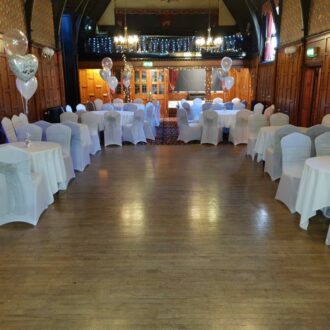 Beautifully dressed dining tables with balloons on top of the tables. The tables are set out around the edge of the room leaving space for a dance floor. The fire place is lit up blue.
