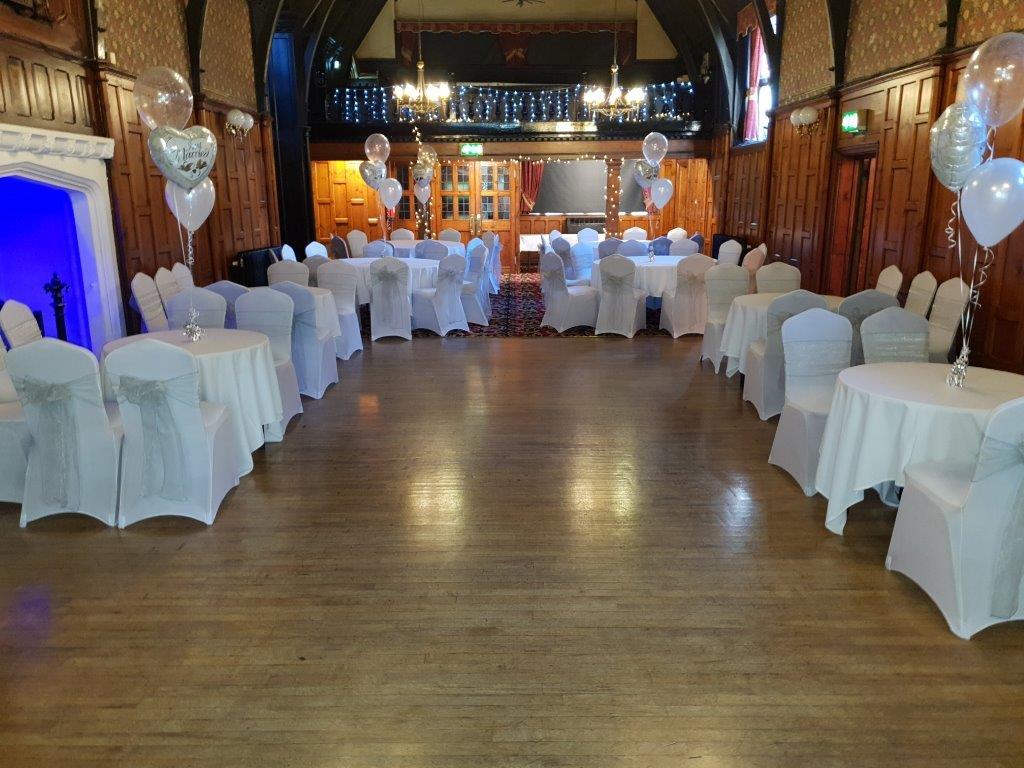 Beautifully dressed dining tables with balloons on top of the tables. The tables are set out around the edge of the room leaving space for a dance floor. The fire place is lit up blue.