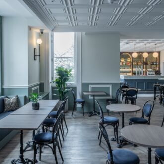 Light-filled café at Buile Hill Mansion featuring wooden tables, bentwood chairs, upholstered bench seating, indoor plants, pale green panelled walls, and a bar visible in the background.