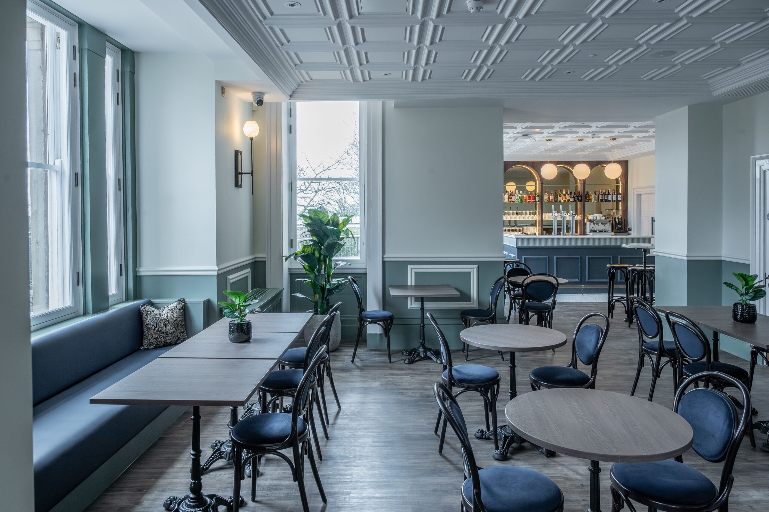 Light-filled café at Buile Hill Mansion featuring wooden tables, bentwood chairs, upholstered bench seating, indoor plants, pale green panelled walls, and a bar visible in the background.